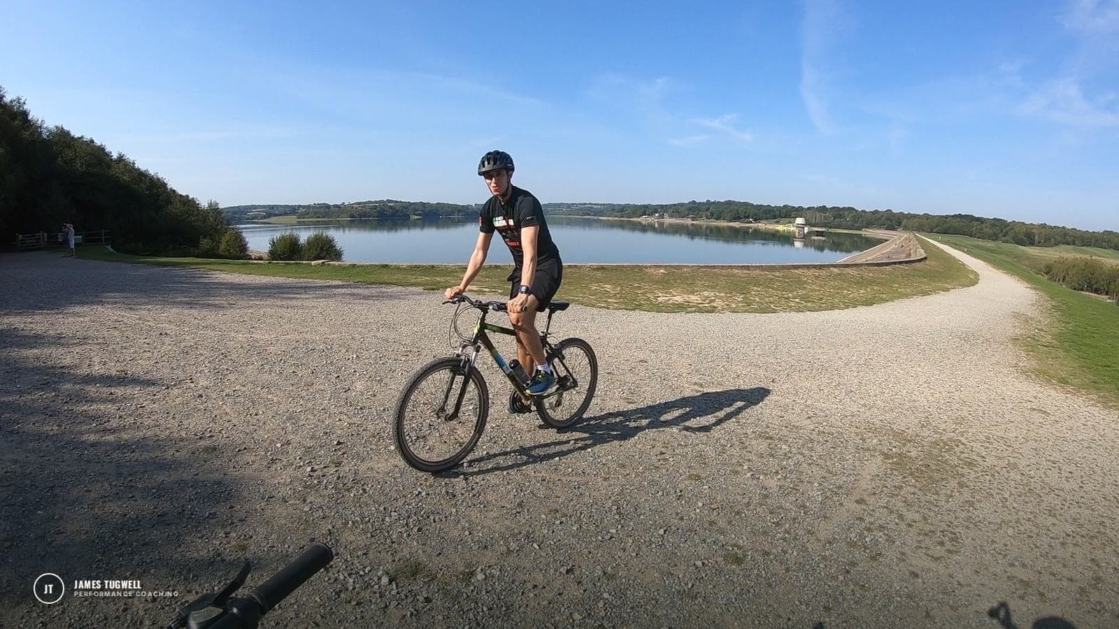James Tugwell riding a bike on a gravel path beside a reservoir on a sunny day